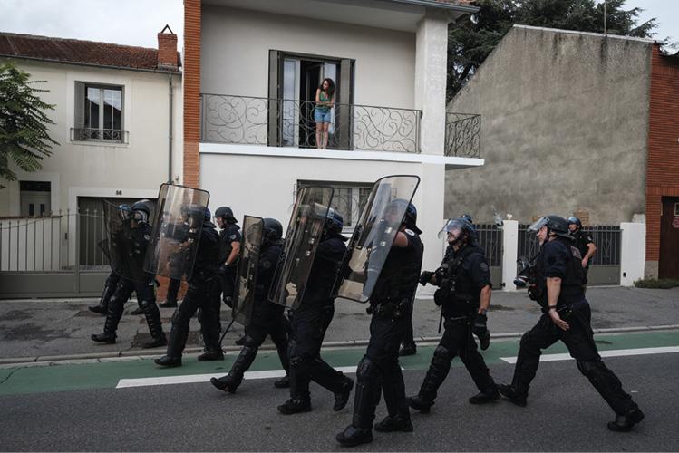 Ranger de flics avancent avec boucliers dressés, dans une rue résidentielle. une femme regarde le hors-champ à gauche depuis son balcon