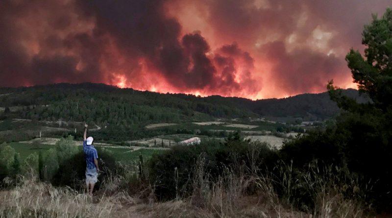 paysage de montagne avec un grand incendie au fond et une personne dos au premier plan avec une casquette blanche à l'envers et la main gauche levée