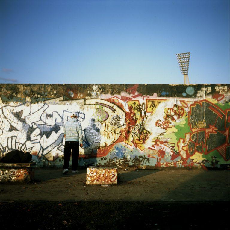d'un mur plein de graffitis, ciel bleu au-dessus et pyl$ône qui dépasse. Une personne de dos.