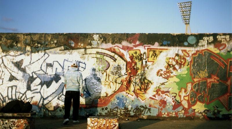 d'un mur plein de graffitis, ciel bleu au-dessus et pyl$ône qui dépasse. Une personne de dos.
