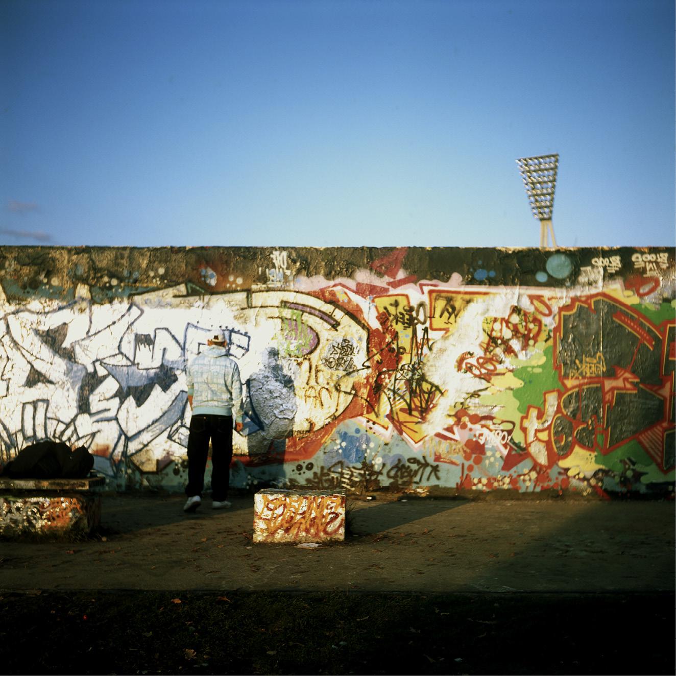d'un mur plein de graffitis, ciel bleu au-dessus et pyl$ône qui dépasse. Une personne de dos.