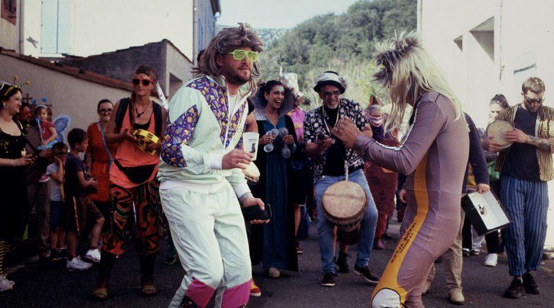 photo de personnes dansant et jouant de la musique dans un village. Le mec au premier plan a des lunettes rayées vert fluo et tient une écocup
