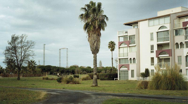 Paysage urbain : à droite, bâtiment résidentiel. Au milieu, palmier. A gauche, structures métalliques hautes et végétation.