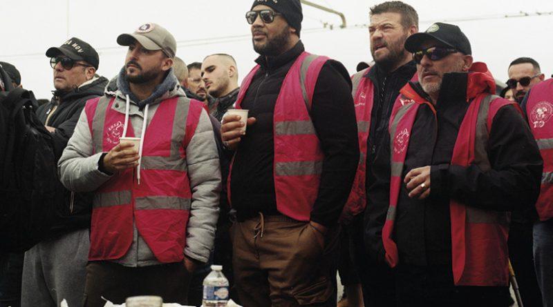 Cinq hommes avec des chasubles de syndicats, regardant vers la gauche. Au premier plan, une table avec baguette, jambon et bouteille d'eau.