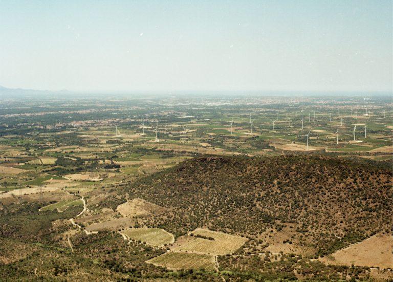 photo du paysage de la plaine du Roussillon. Végétation sèche, beaucoup d'éoliennes, et on distingue à peine une zone habitée (Rivesaltes)