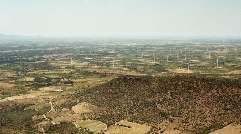 photo du paysage de la plaine du Roussillon. Végétation sèche, beaucoup d'éoliennes, et on distingue à peine une zone habitée (Rivesaltes)