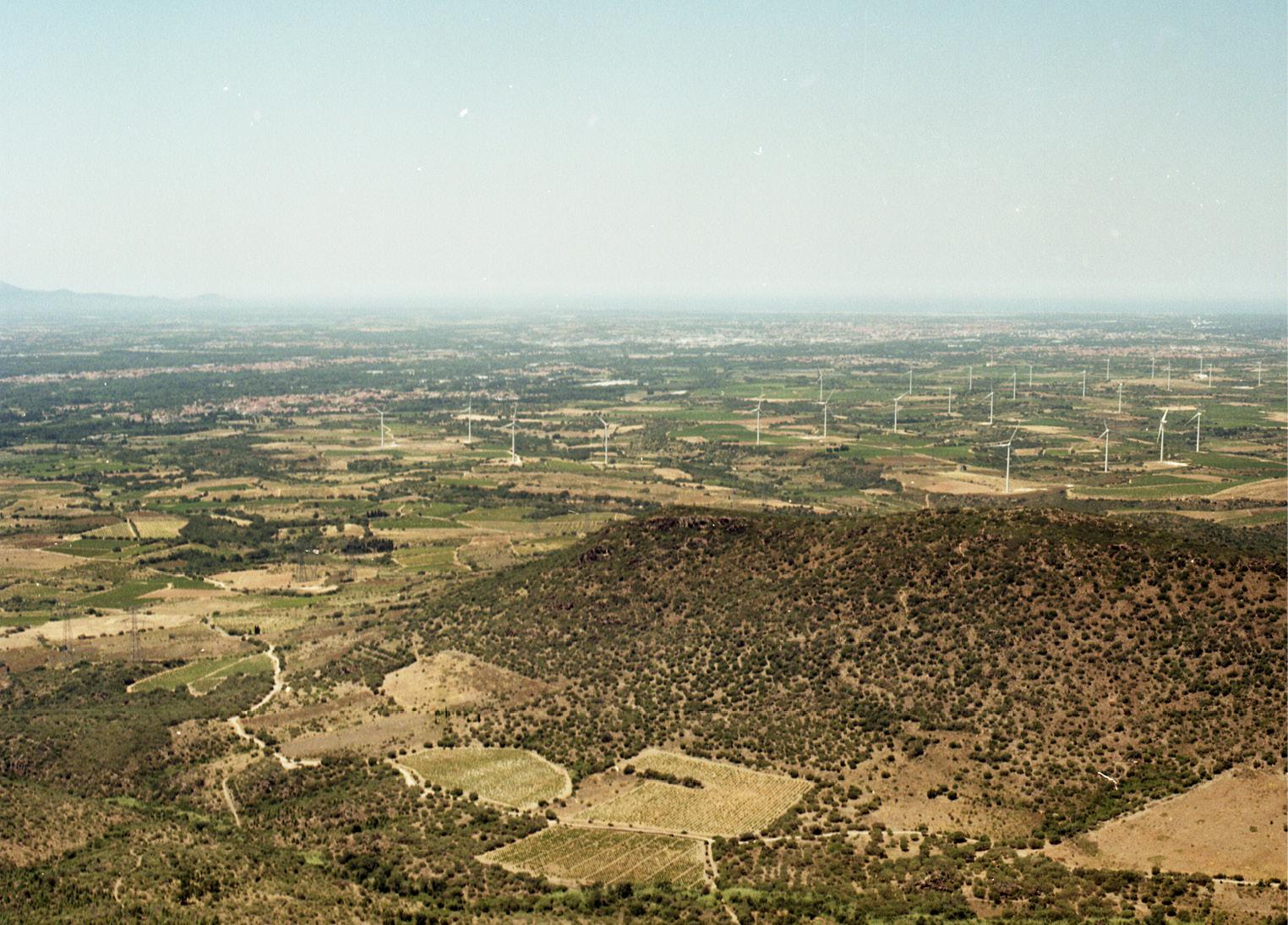 photo du paysage de la plaine du Roussillon. Végétation sèche, beaucoup d'éoliennes, et on distingue à peine une zone habitée (Rivesaltes)
