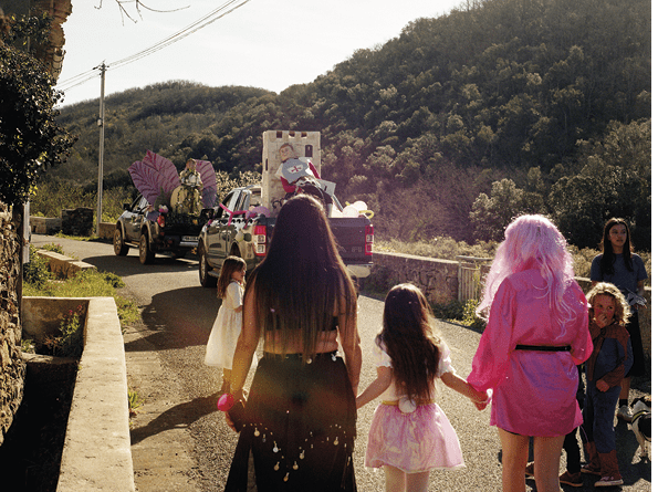 personnes déguisées vues de dos sur une route, voitures avec moniseur carnaval, montagnes