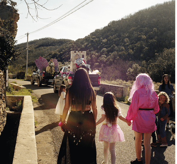 personnes déguisées vues de dos sur une route, voitures avec moniseur carnaval, montagnes