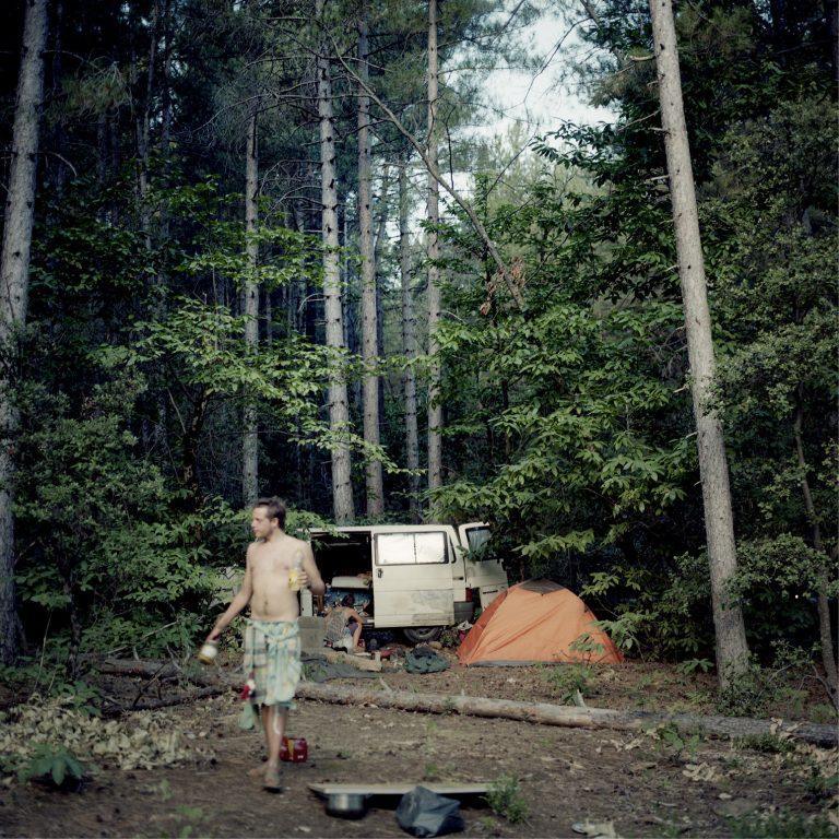 photo. Dans une forêt, un homme, torse nu, en short, a une bouteille a la main. Derrière lui, un camion, une tente, une personne accourpie.