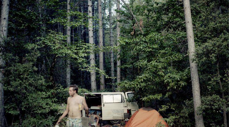 photo. Dans une forêt, un homme, torse nu, en short, a une bouteille a la main. Derrière lui, un camion, une tente, une personne accourpie.